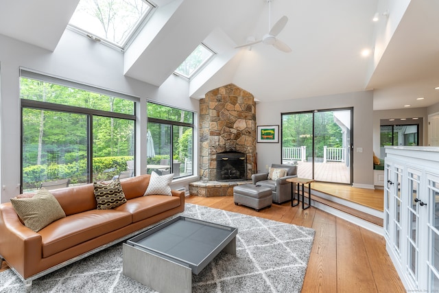 living room with light hardwood / wood-style floors, high vaulted ceiling, a skylight, a stone fireplace, and ceiling fan