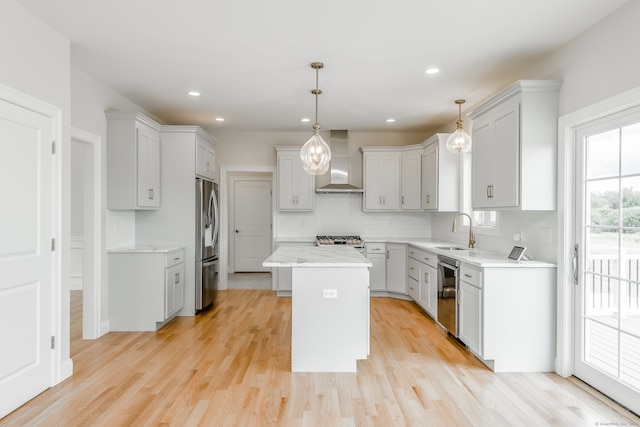 kitchen with appliances with stainless steel finishes, wall chimney range hood, pendant lighting, light hardwood / wood-style floors, and a kitchen island