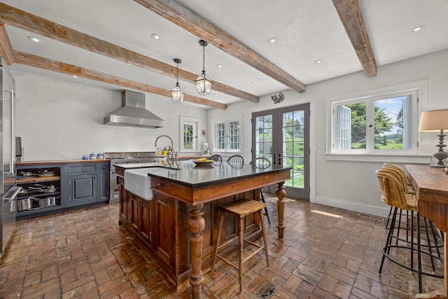 kitchen featuring hanging light fixtures, beam ceiling, wall chimney range hood, and an island with sink