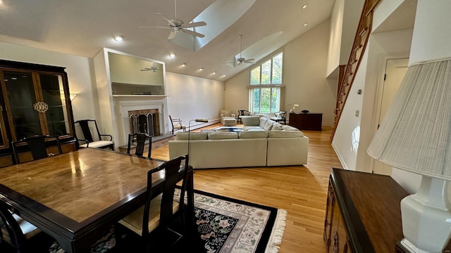 dining room featuring light wood finished floors, a fireplace with raised hearth, high vaulted ceiling, and a ceiling fan