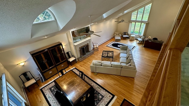 living room featuring high vaulted ceiling, a fireplace with raised hearth, a textured ceiling, wood finished floors, and ceiling fan