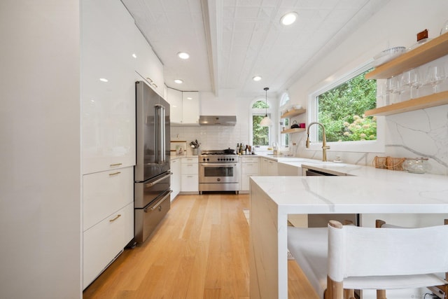kitchen featuring high quality appliances, open shelves, a peninsula, light wood-style floors, and a sink