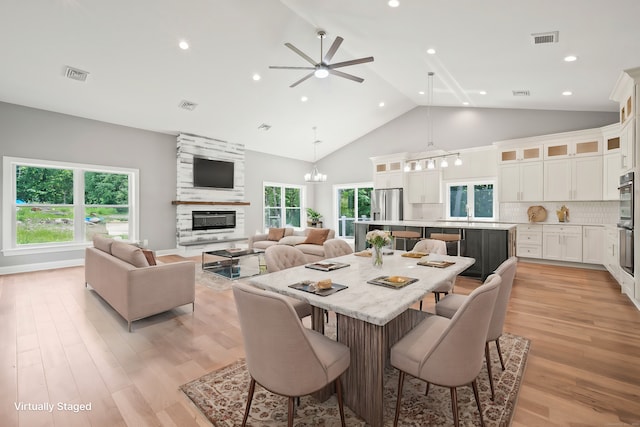 dining room with ceiling fan with notable chandelier, a large fireplace, high vaulted ceiling, and light hardwood / wood-style floors