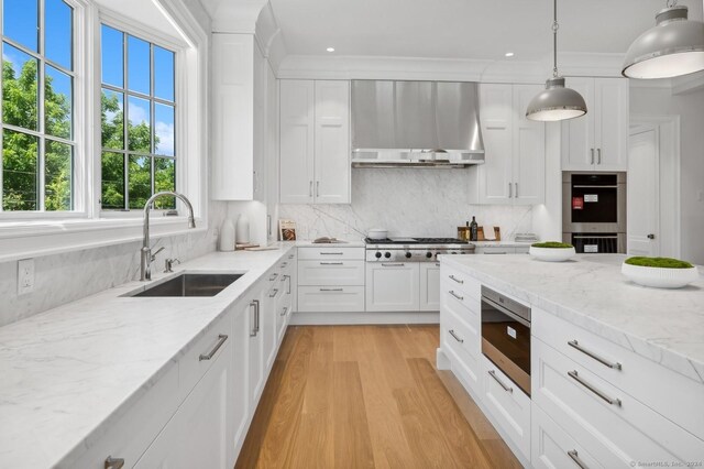 kitchen featuring tasteful backsplash, wall chimney range hood, stainless steel appliances, hanging light fixtures, and light wood-type flooring