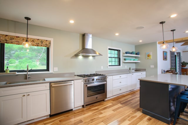 kitchen featuring stainless steel appliances, wall chimney range hood, sink, hanging light fixtures, and white cabinetry