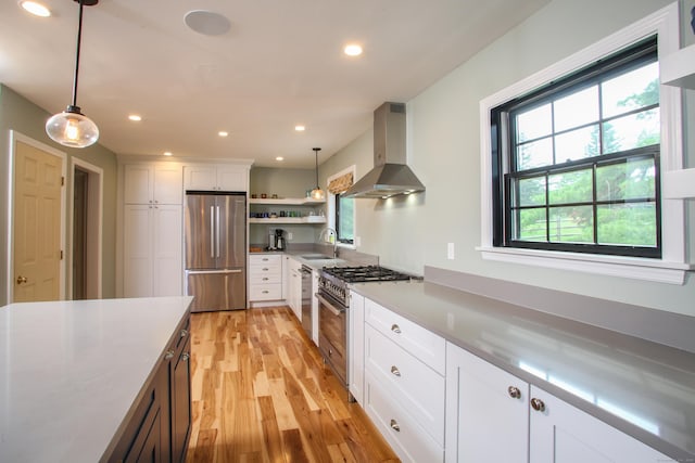 kitchen featuring appliances with stainless steel finishes, white cabinets, wall chimney range hood, pendant lighting, and light wood-type flooring