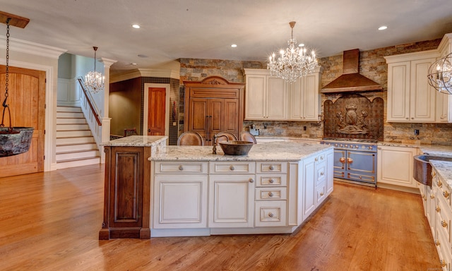 kitchen featuring a kitchen island with sink, wall chimney exhaust hood, light stone countertops, pendant lighting, and light wood-type flooring