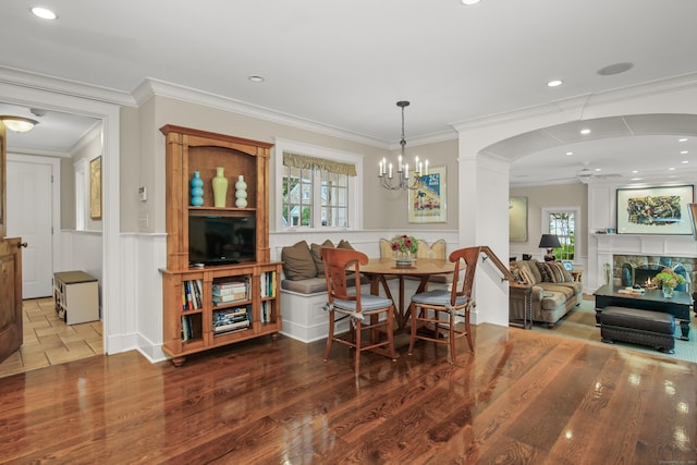 dining area with a wainscoted wall, ceiling fan with notable chandelier, a fireplace, ornamental molding, and dark wood finished floors