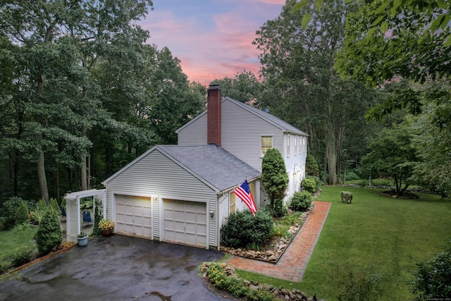 view of side of home with aphalt driveway, a garage, a shingled roof, a yard, and a chimney