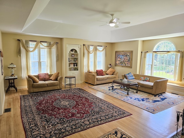 living room with light wood-type flooring, built in features, ceiling fan, and a tray ceiling