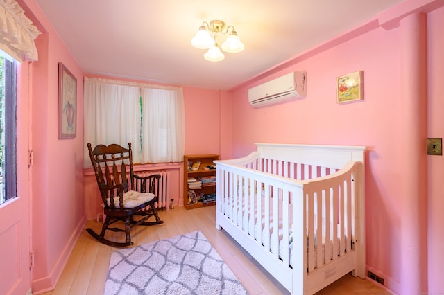 bedroom featuring a crib, an inviting chandelier, a wall mounted air conditioner, and light hardwood / wood-style flooring