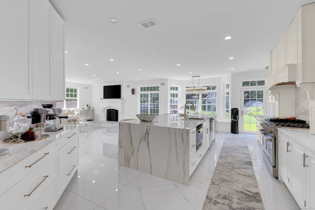 kitchen featuring stainless steel range, backsplash, decorative light fixtures, light stone countertops, and a center island with sink