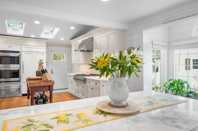 kitchen featuring appliances with stainless steel finishes, a skylight, white cabinets, and wall chimney range hood