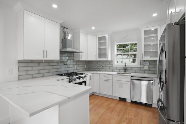 kitchen featuring appliances with stainless steel finishes, white cabinetry, a sink, wall chimney range hood, and a peninsula