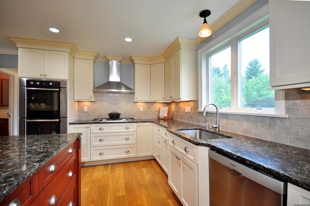 kitchen with stainless steel appliances, dark stone counters, a sink, light wood-type flooring, and wall chimney exhaust hood