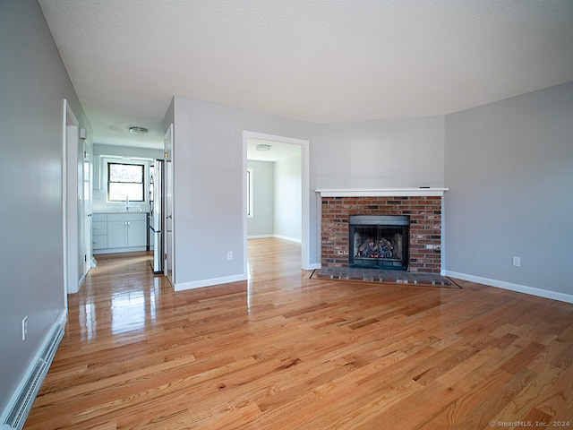 unfurnished living room featuring baseboard heating, light hardwood / wood-style floors, a textured ceiling, and a brick fireplace