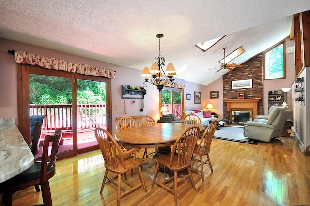 dining space with lofted ceiling with skylight, ceiling fan with notable chandelier, a brick fireplace, light wood-type flooring, and a textured ceiling