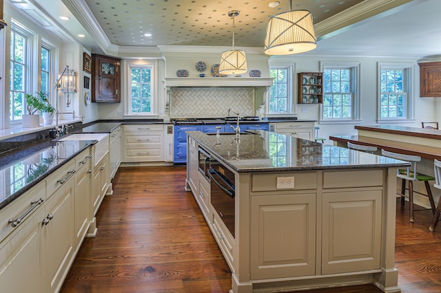 kitchen featuring hanging light fixtures, dark stone countertops, dark wood-type flooring, ornamental molding, and a kitchen island with sink