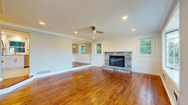 unfurnished living room with visible vents, ornamental molding, wood finished floors, a stone fireplace, and a ceiling fan