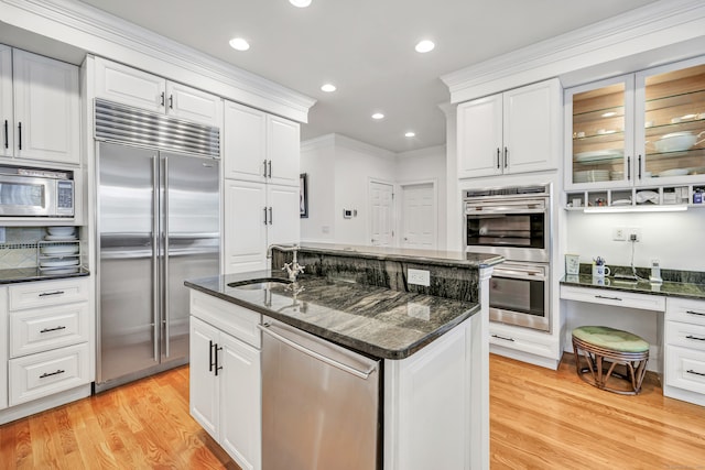 kitchen with white cabinets, a center island with sink, built in appliances, dark stone countertops, and light hardwood / wood-style floors