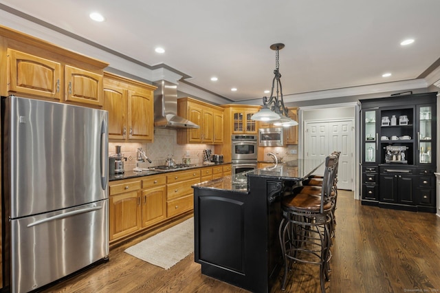 kitchen with wall chimney exhaust hood, a breakfast bar, appliances with stainless steel finishes, dark stone counters, and a kitchen island with sink