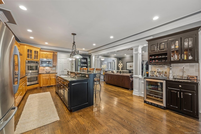 kitchen with sink, appliances with stainless steel finishes, decorative columns, wine cooler, and a center island with sink