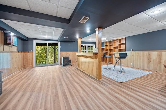 interior space with a wood stove, indoor bar, wooden walls, a drop ceiling, and light hardwood / wood-style floors