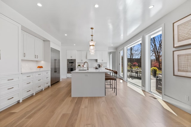 kitchen featuring double oven, a kitchen island with sink, decorative light fixtures, light hardwood / wood-style flooring, and white cabinets