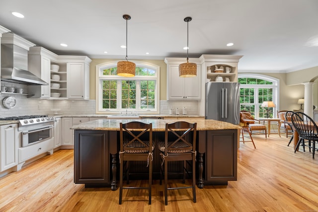 kitchen featuring white cabinets, a kitchen island, wall chimney range hood, stainless steel appliances, and light stone countertops