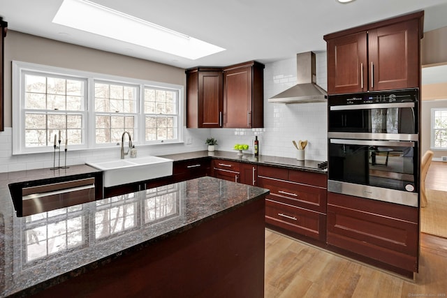 kitchen featuring appliances with stainless steel finishes, tasteful backsplash, dark stone countertops, wall chimney range hood, and light wood-type flooring