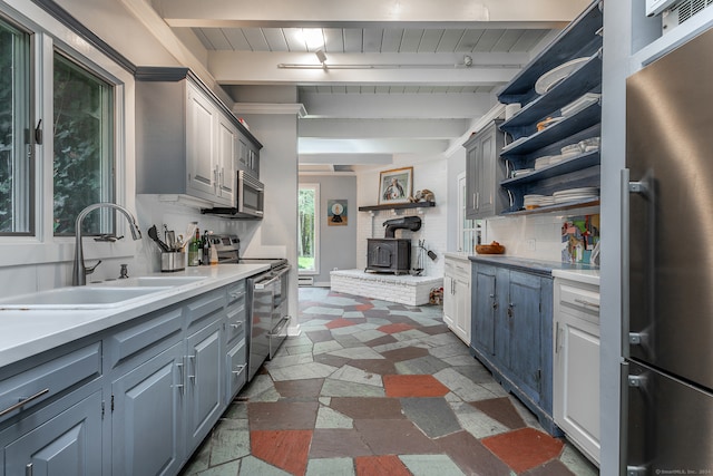 kitchen featuring beamed ceiling, sink, a wood stove, gray cabinets, and appliances with stainless steel finishes