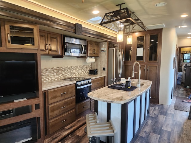 kitchen with an island with sink, stainless steel appliances, glass insert cabinets, tasteful backsplash, and dark wood-style flooring