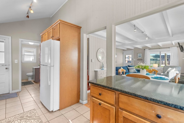 kitchen featuring white refrigerator, dark stone countertops, vaulted ceiling with beams, light tile patterned floors, and track lighting