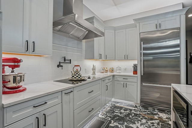 kitchen featuring built in appliances, wall chimney exhaust hood, light stone counters, and tasteful backsplash