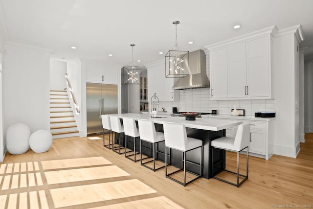 kitchen featuring a kitchen island with sink, wall chimney exhaust hood, light hardwood / wood-style flooring, built in refrigerator, and decorative light fixtures