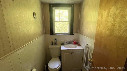 half bath with vanity, toilet, wainscoting, and wallpapered walls