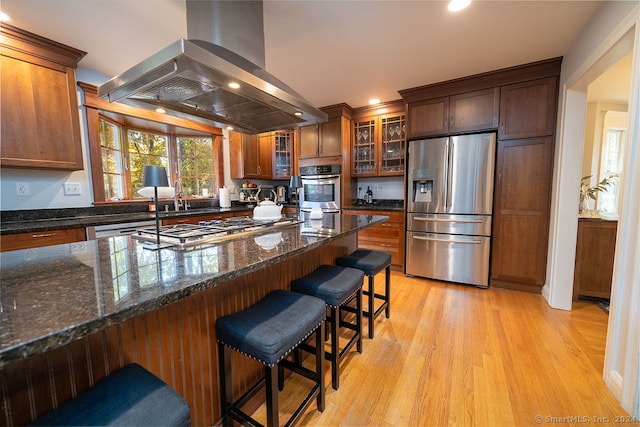 kitchen featuring appliances with stainless steel finishes, light wood-type flooring, island exhaust hood, a kitchen breakfast bar, and dark stone countertops