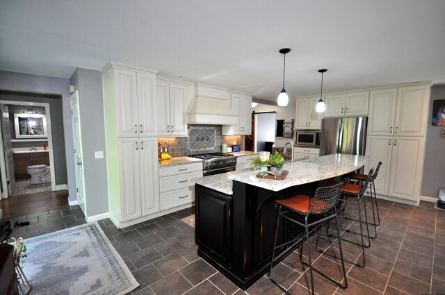 kitchen with custom exhaust hood, light stone counters, an island with sink, stainless steel appliances, and decorative light fixtures