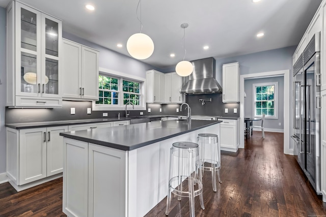 kitchen featuring hanging light fixtures, wall chimney exhaust hood, white cabinets, dark wood-type flooring, and a center island with sink