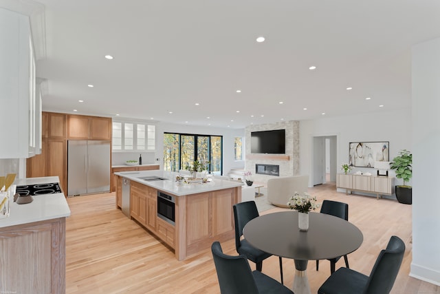 kitchen with light brown cabinets, a large fireplace, a center island with sink, appliances with stainless steel finishes, and light hardwood / wood-style floors