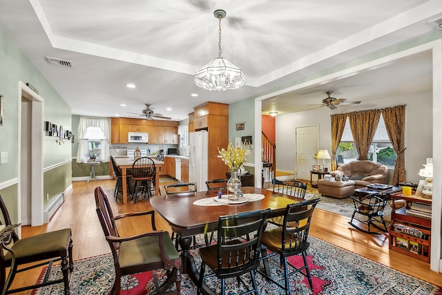 dining room featuring a baseboard radiator, ceiling fan with notable chandelier, a tray ceiling, and light wood-type flooring