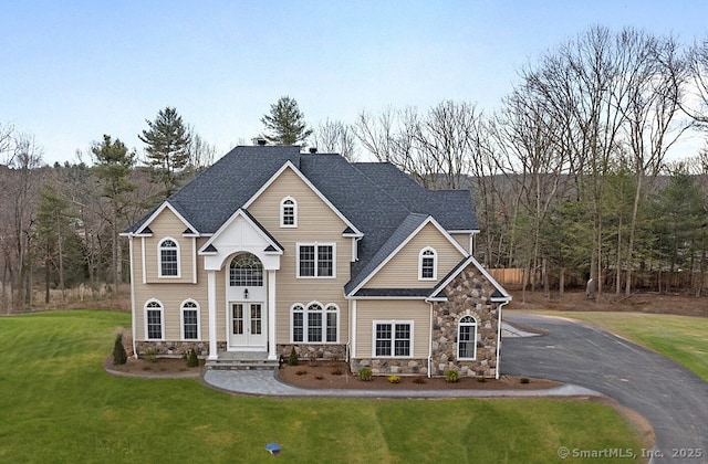 view of front of house with a front lawn, stone siding, driveway, and roof with shingles