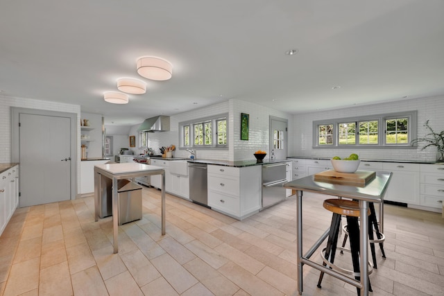kitchen with dishwasher, backsplash, wall chimney exhaust hood, white cabinetry, and dark countertops