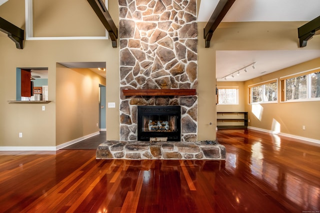 living room with a towering ceiling, a stone fireplace, track lighting, wood finished floors, and baseboards