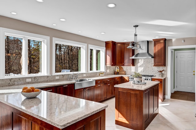 kitchen featuring wall chimney exhaust hood, a center island, light stone counters, and hanging light fixtures