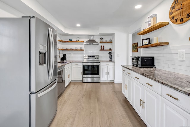 kitchen with dark stone counters, wall chimney range hood, light wood-type flooring, appliances with stainless steel finishes, and white cabinetry