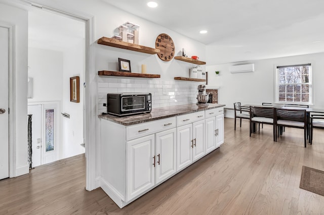 kitchen with white cabinetry, tasteful backsplash, light hardwood / wood-style flooring, a wall unit AC, and dark stone countertops