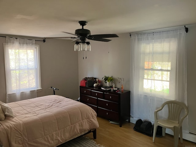 bedroom with ceiling fan, a baseboard heating unit, light hardwood / wood-style flooring, and multiple windows