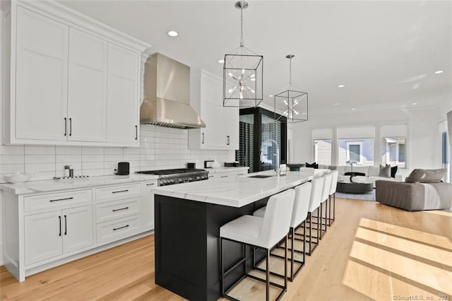 kitchen with light hardwood / wood-style flooring, white cabinetry, hanging light fixtures, a kitchen island with sink, and wall chimney exhaust hood