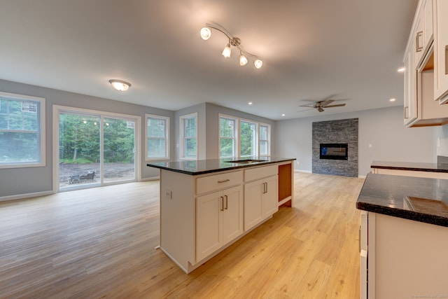 kitchen with white cabinetry, ceiling fan, a stone fireplace, light hardwood / wood-style flooring, and dark stone counters